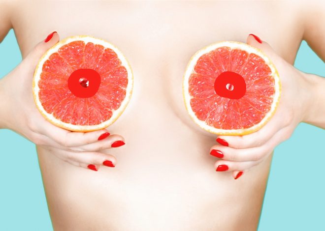 Sexy young woman holding grapefruits near her breasts on color background. Erotic concept